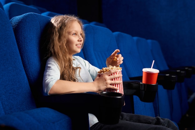 Zijaanzicht van vrouwelijke tiener kijken naar film in lege bioscoop. Meisje dat popcorn eet, rust heeft en in comfortabele stoel ontspant tijdens weekend