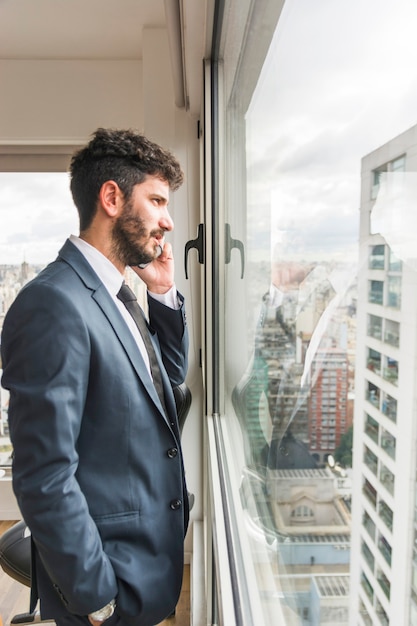 Zijaanzicht van zakenman die zich dichtbij het bureauvenster bevinden die op mobiele telefoon spreken