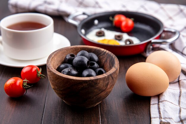 Zijaanzicht van zwarte olijven in kom met de kop thee van eierentomaten op schotelpan van gebraden ei op plaiddoek en houten oppervlakte