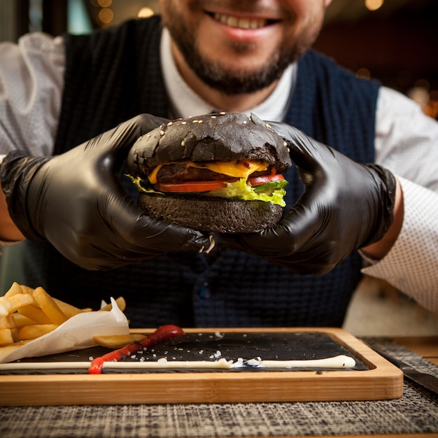 Zijaanzicht zwarte hamburger met wegwerphandschoenen en menselijke hand en frietjes in houten dienblad op restaurant