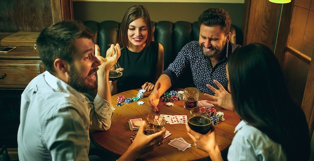 Zijaanzichtfoto van mannelijke en vrouwelijke vrienden die aan houten tafel zitten. mannen en vrouwen die kaartspel spelen. handen met alcoholclose-up.
