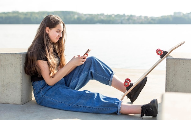 Zijaanzichtmeisje die haar telefoon naast haar skateboard controleren