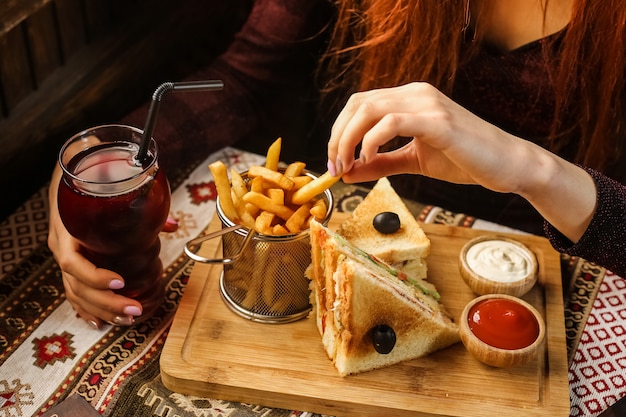 Zijaanzichtvrouw die frieten met dubbeldekkersketchup en mayonaise op tribune met frisdrank eten