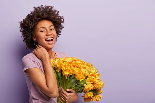 Zijwaarts schot van blije vrouw met donkere huid lacht van vreugde, raakt haar nek, houdt gele tulpen vast, draagt een violet t-shirt, blij met het krijgen van bloemen en complimentjes, poseert over paarse muur, vrije ruimte