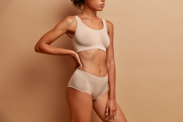 Zijwaarts schot van slanke vrouw draagt cropped top en slipje toont perfect figuur na gewichtsverlies poses tegen beige studiomuur