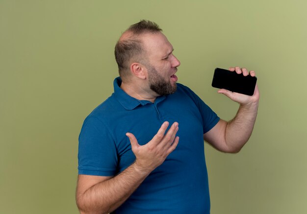 Zingen met gesloten ogen volwassen Slavische man hand in de lucht houden met behulp van mobiele telefoon als microfoon