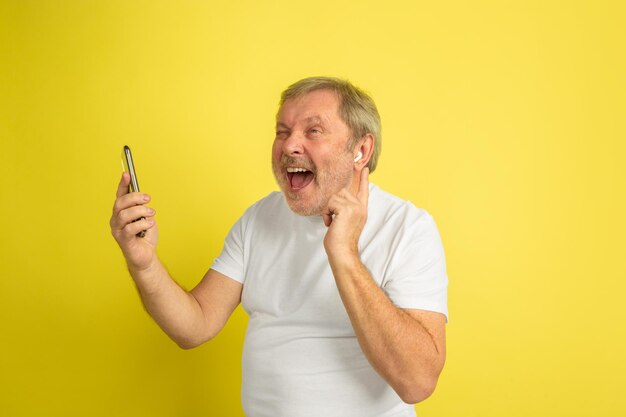 Zingen met oortelefoon en smartphone. Blanke man portret op gele studio achtergrond. Mooi mannelijk model in wit overhemd.
