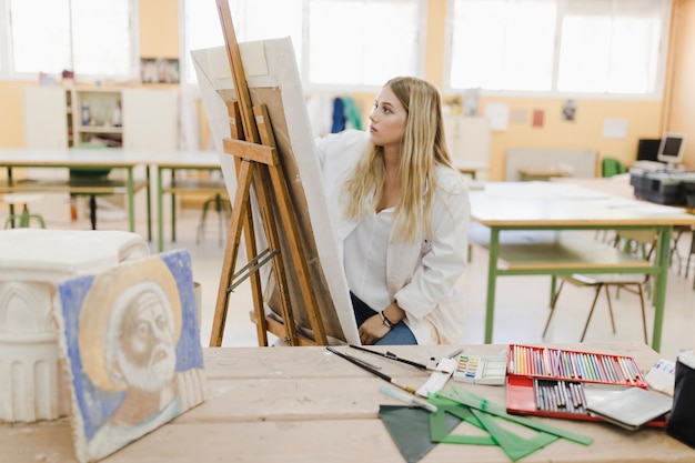 Zitting van de blonde de jonge vrouw in workshop het schilderen op schildersezel