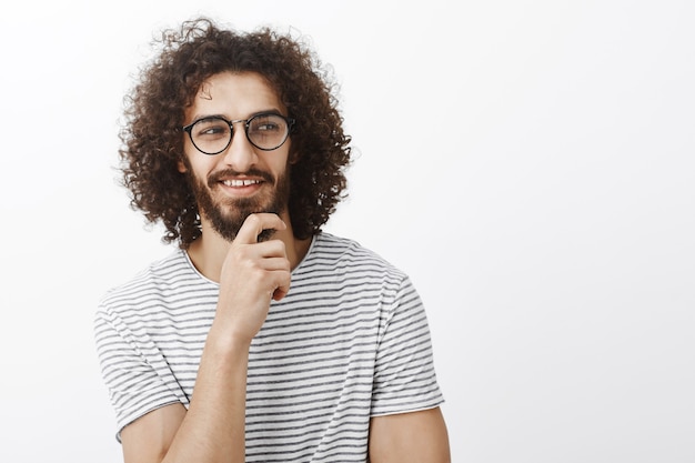 Zoek het uit. Geïntrigeerde aantrekkelijke flirterige man in speelse romantische bui, hand op de baard, er goed uitziend en glimlachend met een tevreden nieuwsgierige uitdrukking