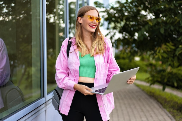 Zomer buitenbeeld van stijlvolle, vrolijke blonde freelance bloggervrouw, die trendy lichte vrijetijdskleding draagt, tas en laptop vasthoudt, klaar voor werk en studie.