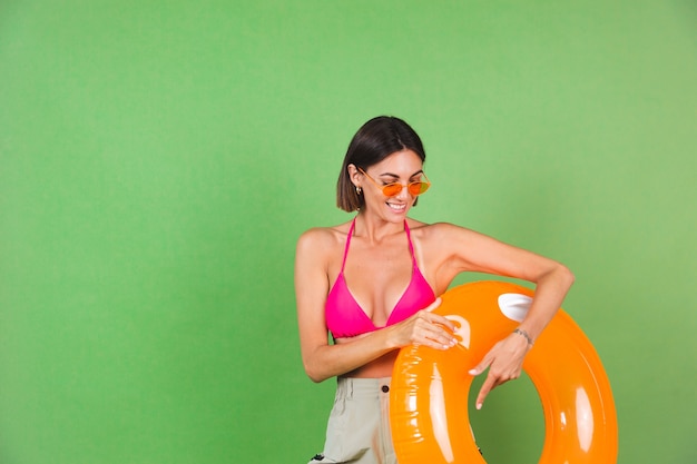 Zomer fit sportieve vrouw in roze bikini en fel oranje opblaasbare ring rond en zonnebril op groen, blij vrolijk opgewonden blij positief