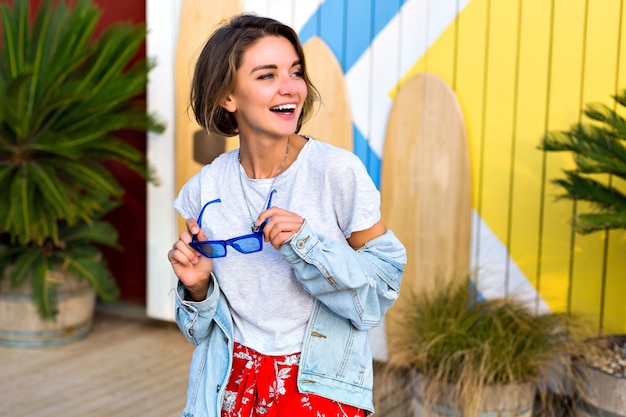 Gratis foto zomer lente heldere positieve portret van gelukkig lachende brunette vrouw trendy vrouwelijke hipster outfit dragen glimlachend en plezier, poseren voor surfplanken en palmen.