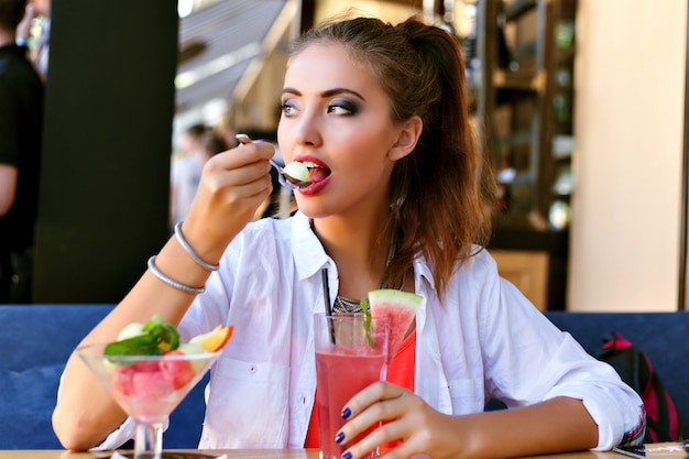 Zomer levensstijl portret van mooie jonge schoonheid vrouw smakelijke woestijn eten op het terras van de stad, geniet van warme dag, verse, sappige cocktail met watermeloen.