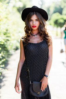 Zomer mode portret van elegante vrouw met perfect golvend haar in stijlvolle elegante zwarte hoed en lichte make-up poseren in het park.