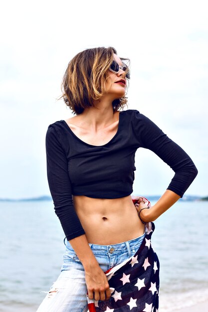 Zomer mode portret van prachtige fit sexy vrouw, gekleed in denim en crop top, met Amerikaanse vlag, tijd doorbrengen op het strand op een regenachtige dag.