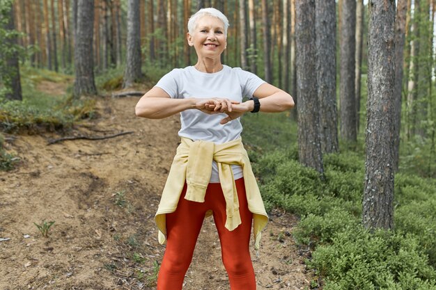 Zomer-, sport-, activiteits- en welzijnsconcept. Mooie energieke gepensioneerde vrouw buitenshuis trainen, lichaam voorbereiden op run, opwarmen, spieren strekken, staande op pad tussen bomen