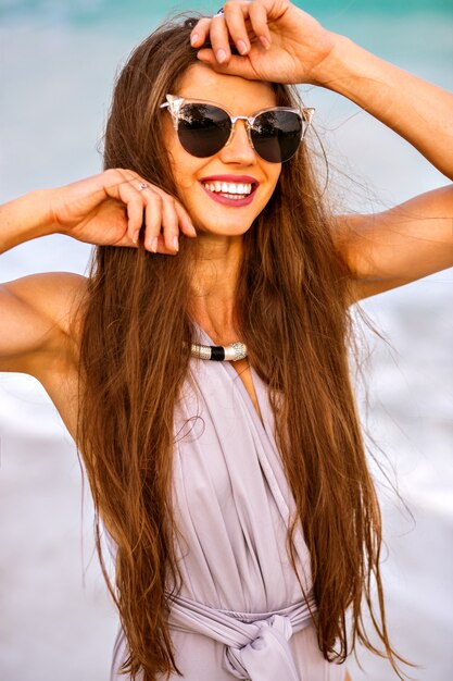 Zomer strand portret van mooie brunette vrouw met sportieve perfect gebruinde lichaam en lange donkerbruine haren, trendy glamour elegante zwembroek dragen, model ontspannen in de buurt van oceaan, zonnebril en sieraden.