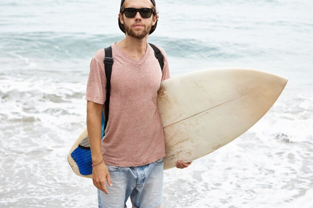 Zomervakanties, actieve levensstijl en vrije tijd concept. Openluchtportret van jonge surfer in zwarte schaduwen die witte surfplank onder zijn wapen en het kijken houden