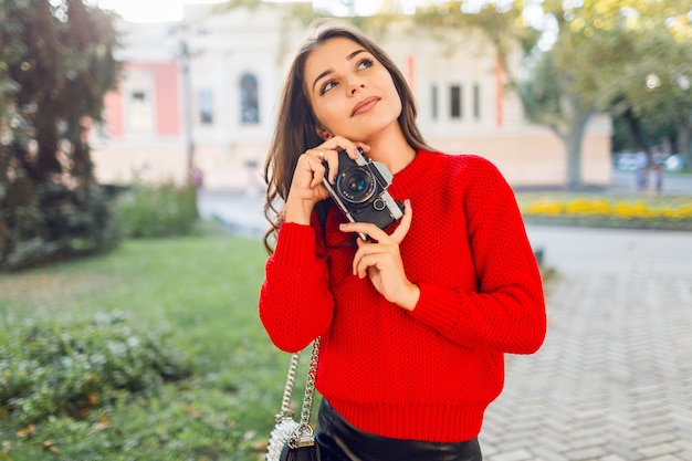 Zonnig levensstijlbeeld van vrij donkerbruin meisje in rode toevallige trui en rok die foto's maken door fotocamera in zonnig park. Wandelen in de stadstuin en op zoek naar bezienswaardigheden.