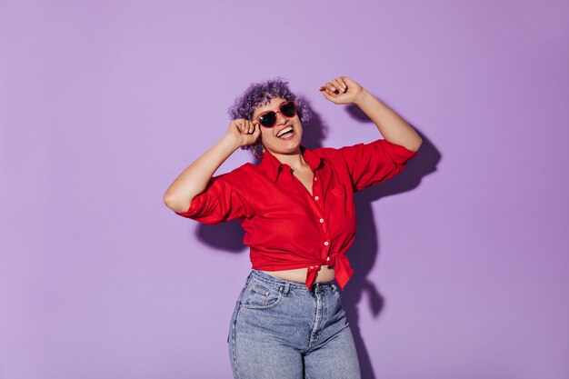 Zonnige heldere vrouw in rood shirt met lange mouwen vastgebonden in de taille. Kortharige vrouw met lila haar in jeans en glazen glimlachen.