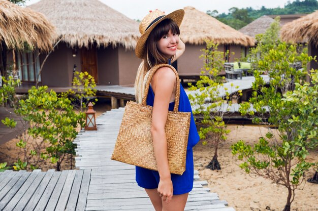 Zonnige zomer portret van schattige brunette vrouw in strooien hoed en trendy boho tas wandelen in tropische villa in Thailand.