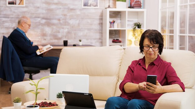 Zoom in op een foto van een oudere vrouw met een bril die een slokje koffie neemt terwijl ze aan het browsen is op de telefoon. Oudere leeftijd man leest een boek op de achtergrond.