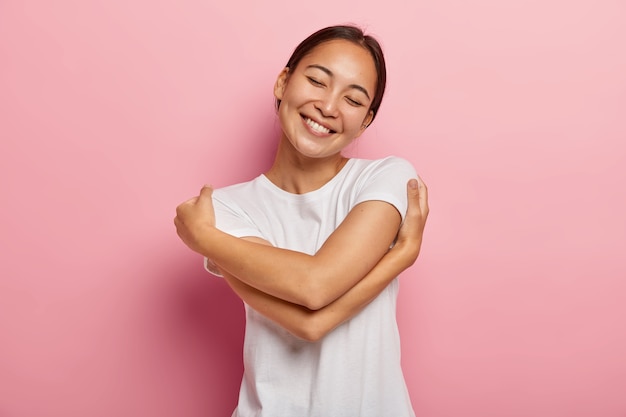 Zorg en tederheid concept. Vrolijke positieve brunette Aziatische vrouw knuffelt zichzelf zachtjes, kantelt het hoofd en lacht vrolijk, houdt de ogen dicht van plezier, heeft een romantische bui, gekleed in een casual outfit