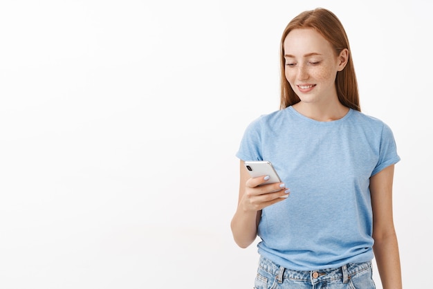 Zorgeloos blij en vermaakt aantrekkelijke roodharige vrouw met sproeten in blauw t-shirt sms'en via nieuwe smartphone verheugd staren en apparaatscherm