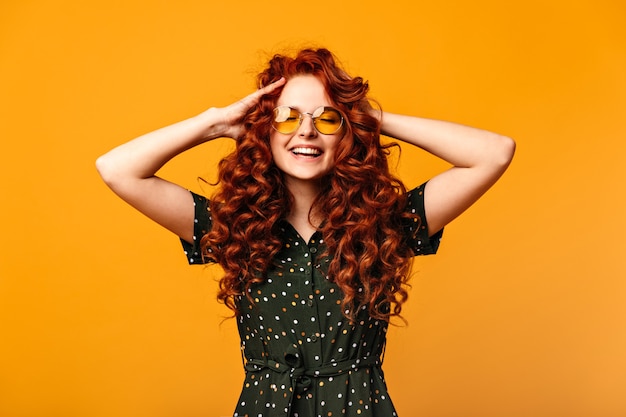 Zorgeloos gembermeisje dat met gesloten ogen lacht. Studio shot van extatische jonge vrouw aanraken van krullend haar op gele achtergrond.