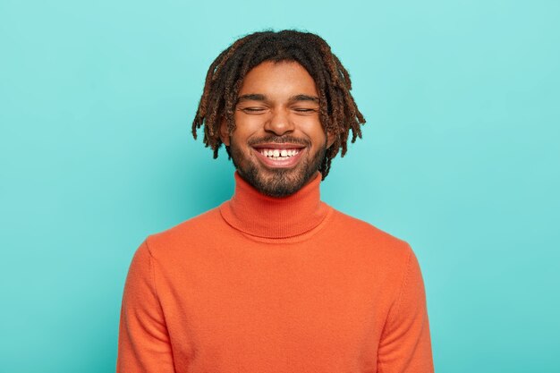 Zorgeloos glimlachende man heeft een vrolijke gezichtsuitdrukking, lacht om iets positiefs, toont witte tanden, draagt een oranje poloneck