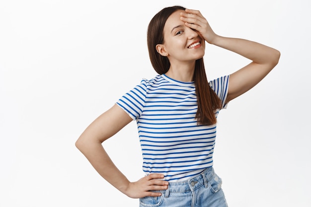 Zorgeloos lachende vrouw bedekt één oog met de hand, kijkt gelukkig naar de voorkant, houdt de hand op de taille, draagt een gestreept zomert-shirt, staat over een witte muur