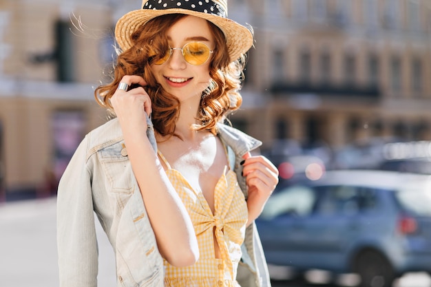 Zorgeloos meisje in zonnebril en spijkerjasje genieten van warme lentedag. Buiten schot van prachtige gember jonge vrouw die zachtjes glimlacht tijdens wandeling.