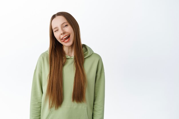 Zorgeloos mooi millennial meisje toont tong, glimlachend met witte perfecte tanden en knipogend, staande in hoodie met positieve emotie, studiomuur