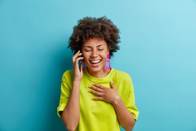 Zorgeloos ontspannen positief Afro-Amerikaanse vrouw lacht uit terwijl praat via smartphone hand op borst houdt ogen sluit positieve emoties hoort grappige grap terloops gekleed staat binnen