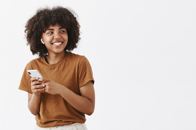 zorgeloos vriendelijk en creatief stijlvol Afrikaans Amerikaans tienermeisje in bruin t-shirt draait rechtsaf met brede tevreden glimlach met draadloze koptelefoon en smartphone vast te houden