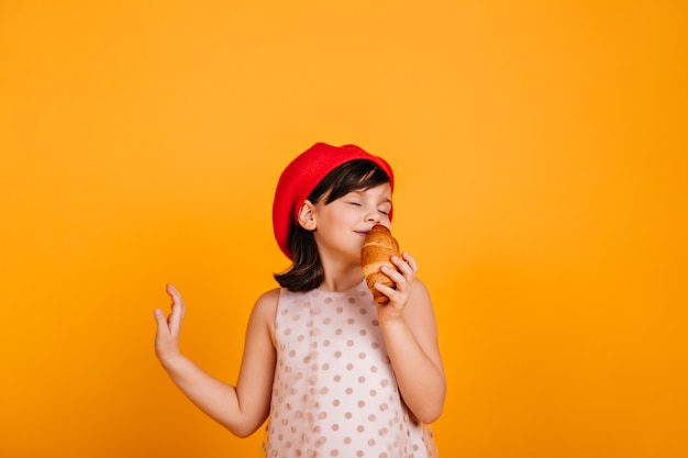 Zorgeloos vrouwelijk kind dat croissant eet. schattig jong geitje dat zich op gele muur bevindt.