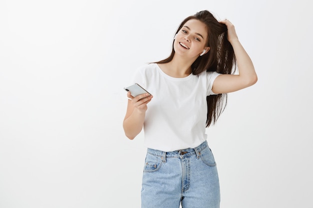 Zorgeloos zelfverzekerd jonge vrouw poseren met haar telefoon en oordopjes tegen witte muur