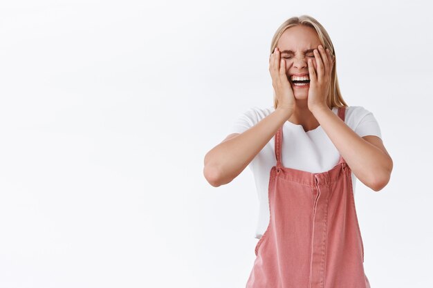 Zorgeloze, extraverte, knappe blanke blonde vrouw in casual outfit, knijp in de wangen en lach hard als kijken naar grappige komische film, plezier maken, gek doen met vrienden, grinniken, witte achtergrond