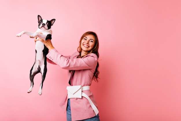 Zorgeloze jonge dame met zwarte franse bulldog met oprechte glimlach. indoor portret van vrolijk meisje spelen met hond op pastel.