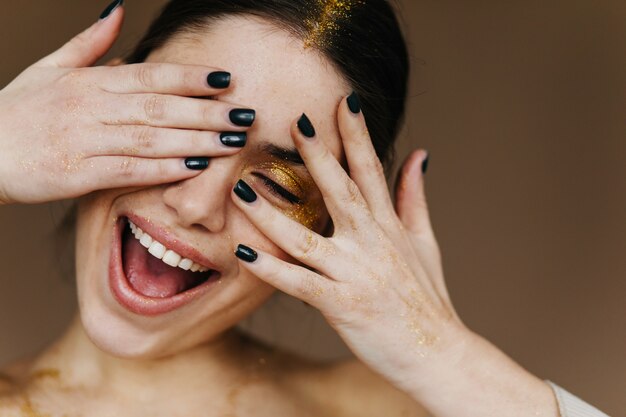 Zorgeloze jonge vrouw die met partijmake-up geluk uitdrukt. geïnspireerd donkerbruin meisje dat op donkere muur lacht.