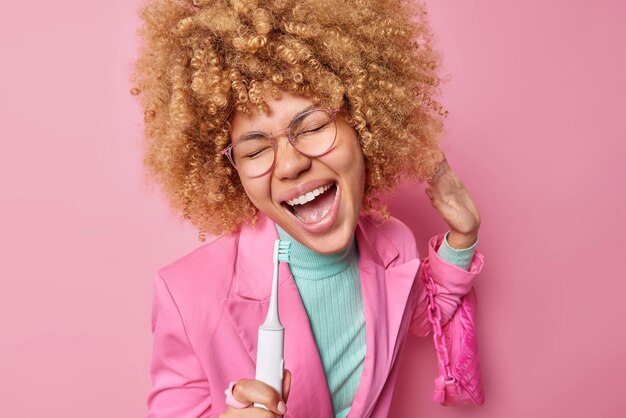 Zorgeloze optimistische vrouw heeft plezier tijdens het tandenpoetsen houdt elektrische tandenborstel in de buurt van mond alsof microfoon favoriete liedje zingt draagt damestas gekleed in stijlvolle kleding geïsoleerd over roze muur