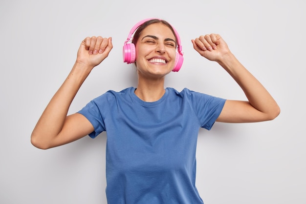 Zorgeloze positieve vrouw schudt armen dansen met ritme van muziek houdt ogen gesloten glimlacht graag stereo hoofdtelefoon en casual blauw t-shirt geïsoleerd op witte achtergrond heeft een gelukkige stemming