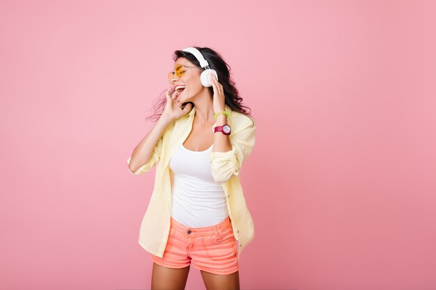 Zorgeloze Spaanse vrouw in roze korte broek zingen terwijl poseren. Indoor portret van modieus Aziatisch meisje in witte tank-top toucnhing hoofdtelefoons en rondkijken.