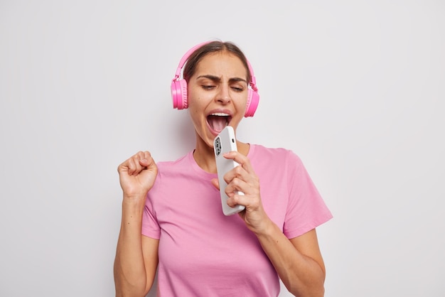 Zorgeloze vrolijke vrouw zingt lied houdt smartphone in de buurt van mond alsof microfoon plezier heeft dwazen rond gekleed in casual roze t-shirt geïsoleerd op witte achtergrond Muziek en entertainment