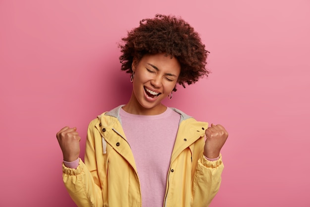 Zorgeloze zwarte vrouw balt vuisten van vreugde, kantelt het hoofd en voelt zich als winnaar, viert de overwinning, sluit de ogen met plezier, draagt casual trui en jas