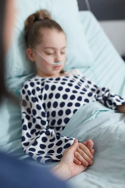 Zorgzame moeder die de hand van de zieke dochter vasthoudt terwijl ze ontspannen op het bed van de pediatrische kliniek ligt. verdoofd jong meisje dat een voorzichtige vrouwenhand vasthoudt terwijl ze slaapt in de herstelafdeling van het ziekenhuis.