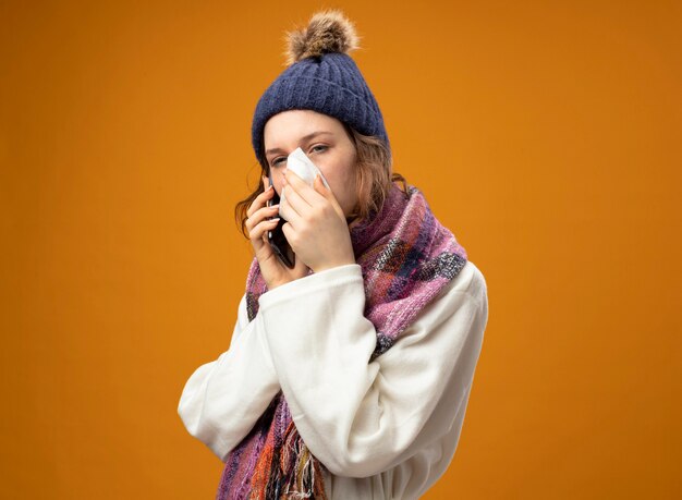 Zwak jong ziek meisje kijken kant dragen witte mantel en winter hoed met sjaal spreekt op telefoon neus afvegen met servet geïsoleerd op oranje