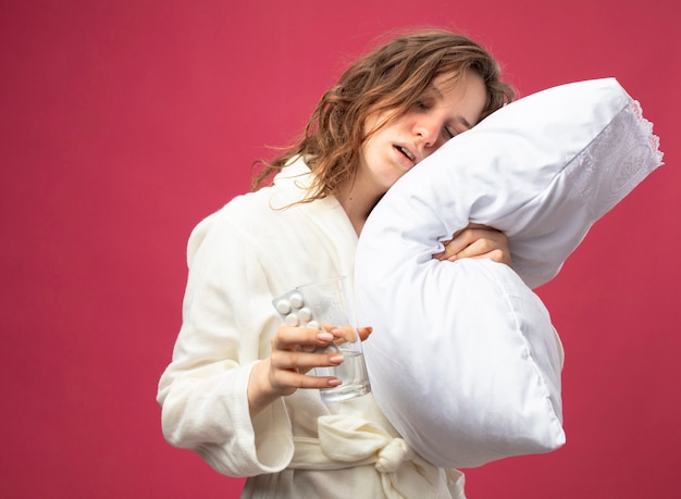 Gratis foto zwak jong ziek meisje met wit gewaad omhelsde kussen met glas water