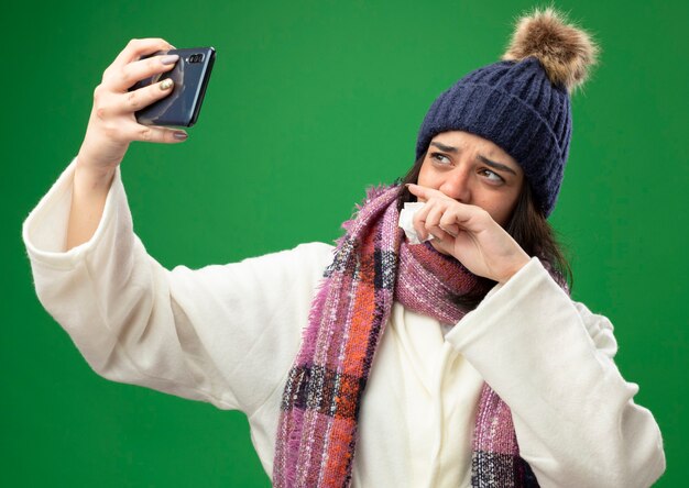 Zwakke jonge kaukasische ziek meisje dragen gewaad winter muts en sjaal houden servet hand houden op mond nemen selfie geïsoleerd op groene achtergrond