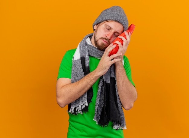 Zwakke jonge knappe slavische zieke man met winter hoed en sjaal met warm waterzak aanraken van gezicht met het met gesloten ogen geïsoleerd op een oranje achtergrond met kopie ruimte